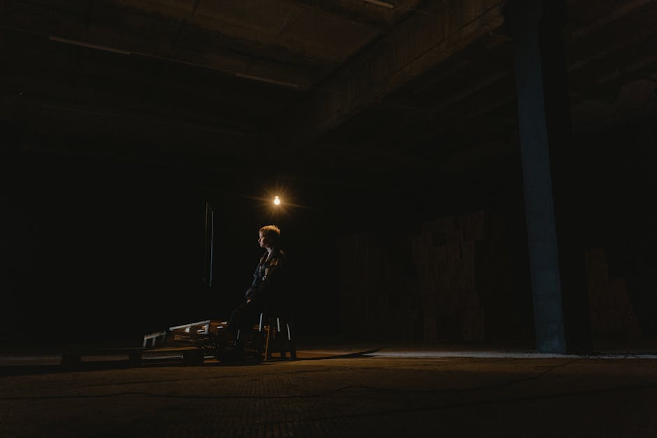 A solitary figure seated in a dark industrial space, illuminated by soft light.