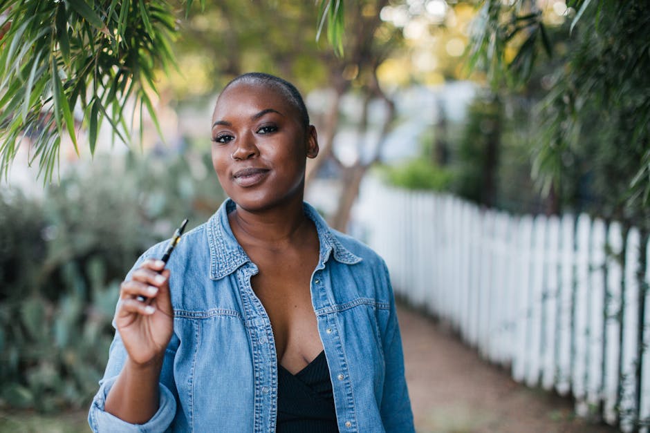 Stylish African American woman outdoors in a denim jacket holding a vape pen.