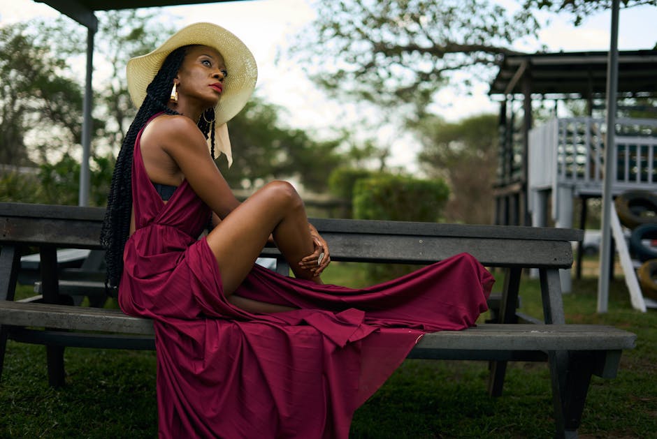 Stylish woman in a flowing burgundy dress and sun hat sitting on an outdoor bench.