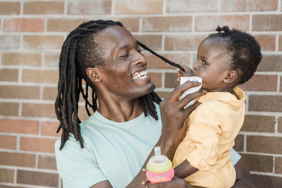 Smiling father with dreadlocks lovingly engages with his baby daughter outdoors against a brick wall.