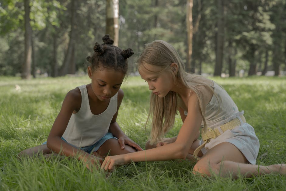 Two girls creating bead necklaces while sitting on grass in a sunny park.