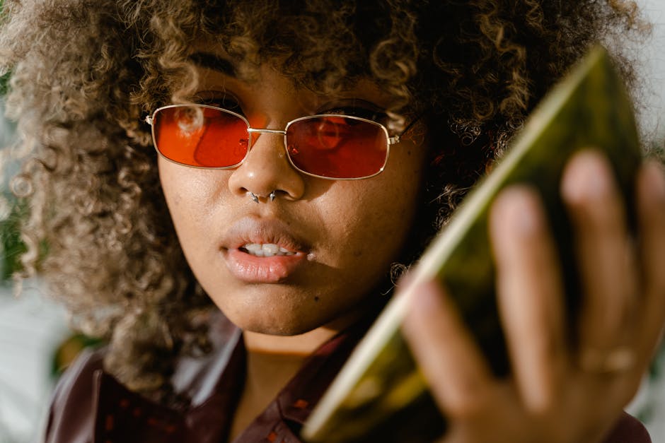 Confident young woman with afro hair and red sunglasses holding a watermelon slice outdoors.