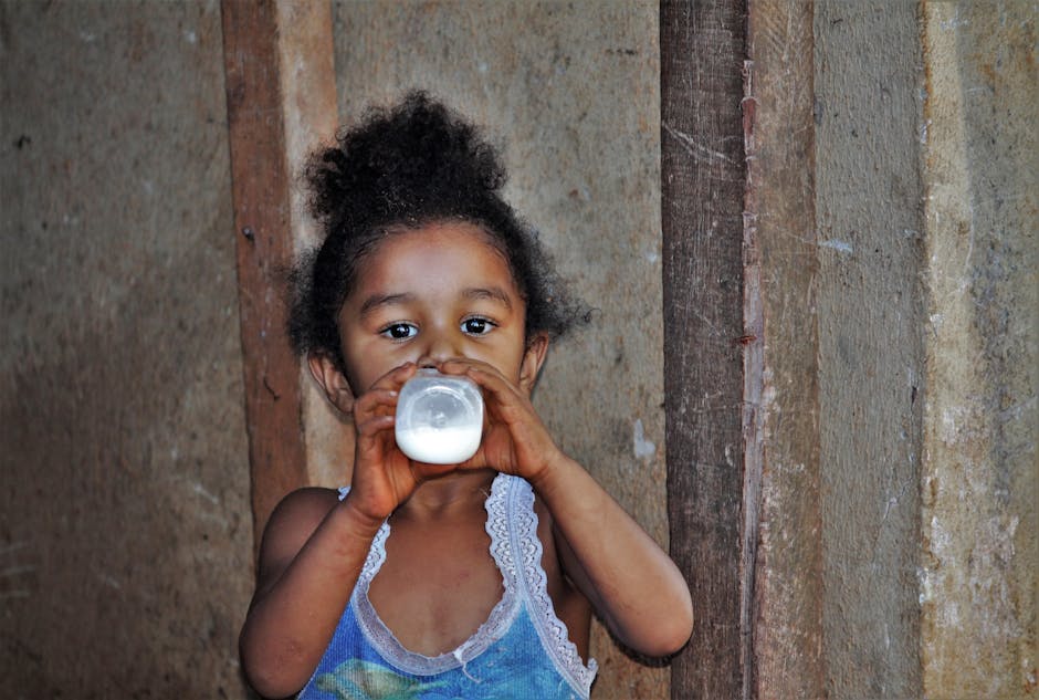 A young girl with curly hair drinks milk from a bottle against a wooden background.