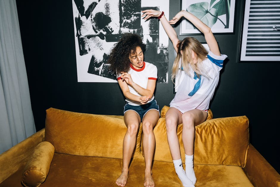 Two young women joyfully dancing together on a yellow sofa indoors.