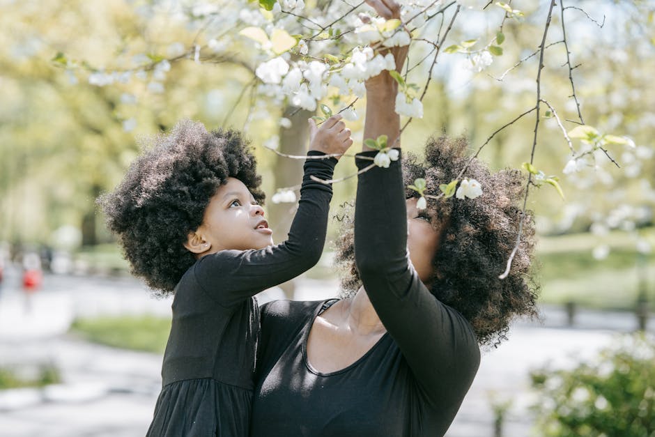 A mother and daughter picking white blossoms together in a sunny outdoor park.