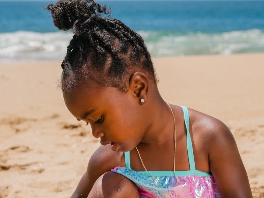 A young girl with braided hair is engrossed in play on a sunny beach day, wearing colorful swimwear.