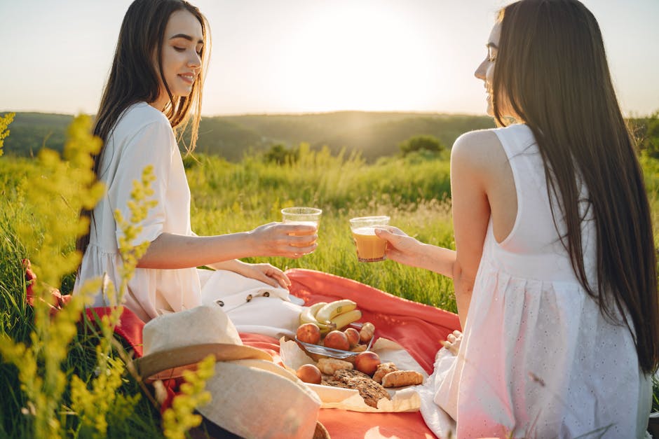Two women enjoying a summer picnic with fresh fruit and juice in a scenic field.
