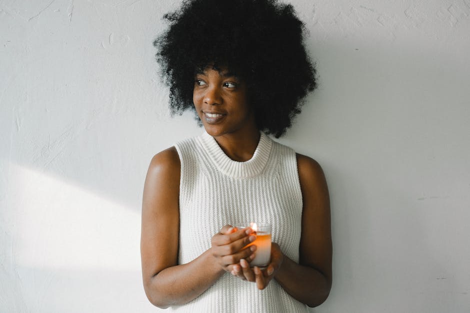 Gentle African American female with curly hair standing with aromatic burning candle in glass at home and looking away