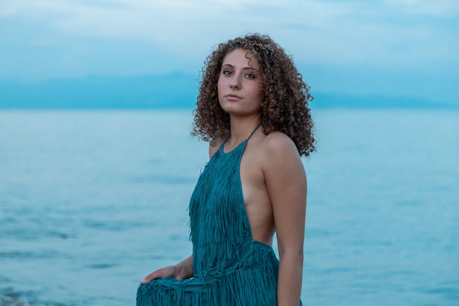 A beautiful woman with curly hair posing in a blue dress by the sea during the day.