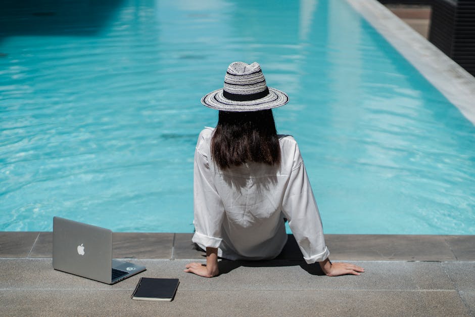 Young woman in white shirt working remotely by the pool, enjoying a sunny summer day.