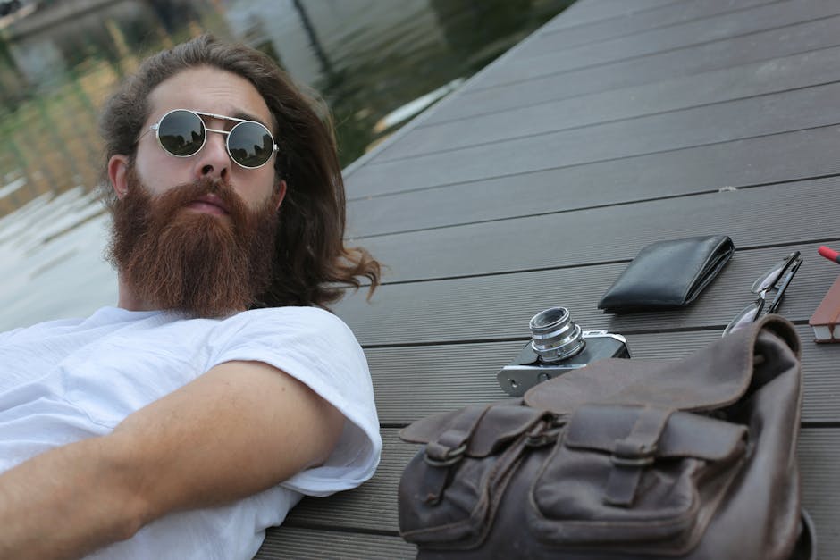 A stylish young man with a beard and sunglasses relaxes by the water, accompanied by a vintage camera and leather bag.
