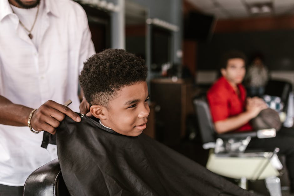 A young boy happily getting a haircut inside a modern barbershop, observed by an adult.