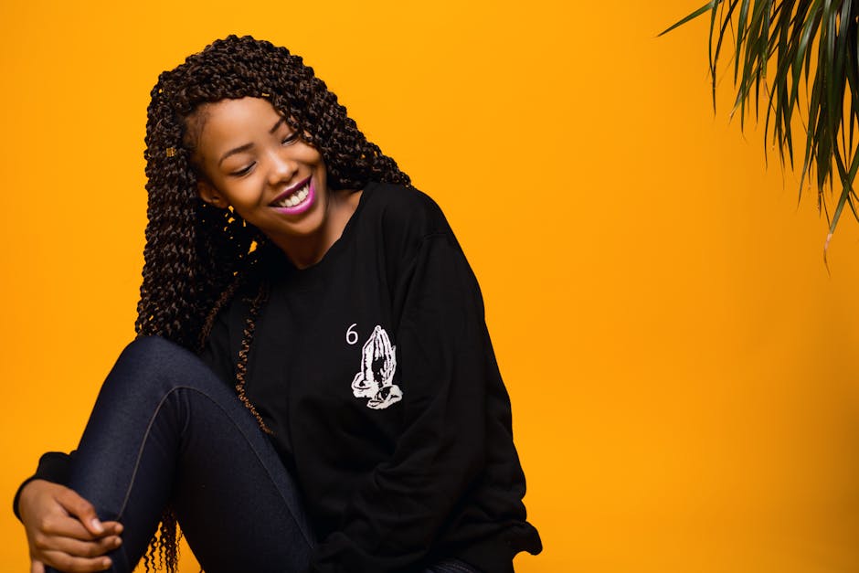Happy smiling young African American lady with long Afro hair embracing knee and looking away against yellow background